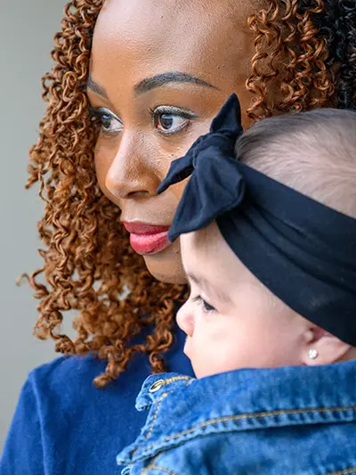 A woman with brown curly hair and red lipstick holds a baby wearing a denim jacket and black bow headband.
